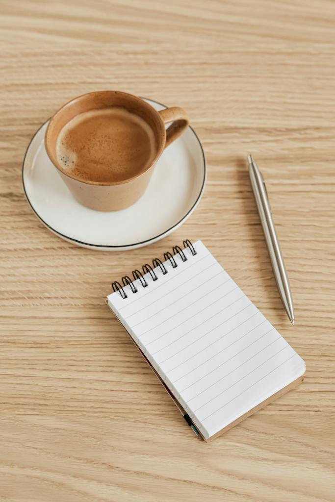 From above of opened notebook with blank sheet near silver pen composed with fragrant cup of hot espresso placed on wooden table